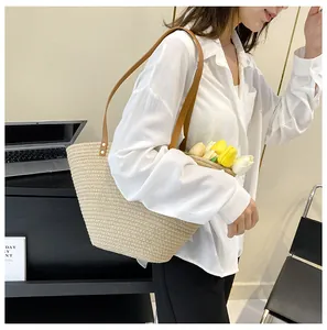Large beige woven straw tote bag with brown leather shoulder straps worn by model