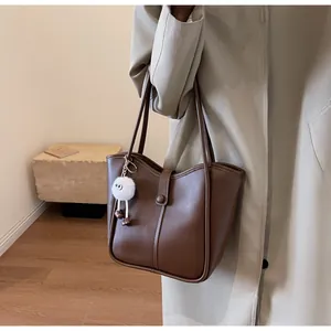 Detailed side view of a brown leather tote bag with a white plush charm on a beige coat