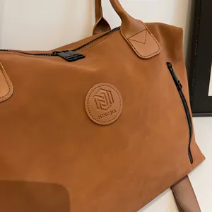 Macro detail of brown travel bag showing suede texture, metal zipper and leather logo patch