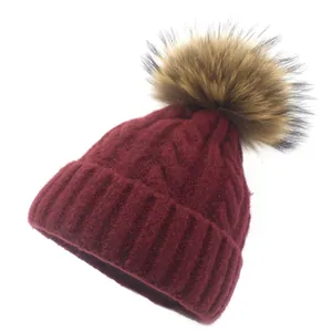 Detail of burgundy knit beanie featuring chunky cable patterns and a large faux fur pom-pom