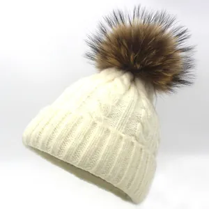 Close-up of beige knit beanie featuring thick cable patterns and large raccoon fur pom-pom