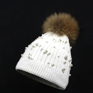 Close-up detail of white knitted beanie featuring luxury pearls and a brown faux fur pompom