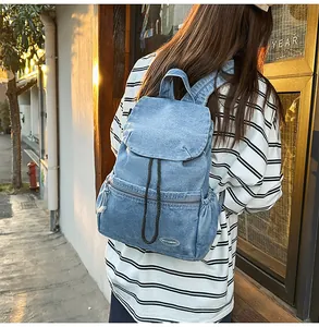 Outdoor back view of a light blue washed denim backpack with drawstring flap and zip pocket