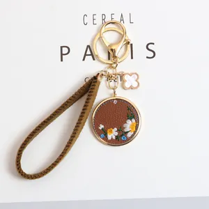 Detail of embroidered daisy keychain with leather charm and clover gold pendant