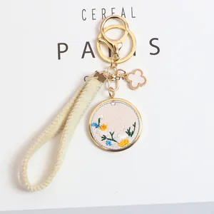 Detail of embroidered daisy round pendant and beige woven strap car keychain with gold hardware