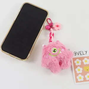 Pink plush monster keychain with bell and flower accessory next to phone for size scale