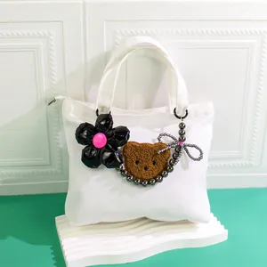 Detail of white canvas tote bag with black flower, plush bear charm, and beaded chain decor