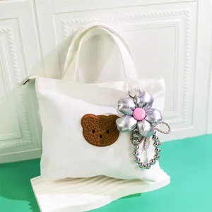 White canvas tote bag featuring a plush bear patch and silver floral charm on a wave pedestal
