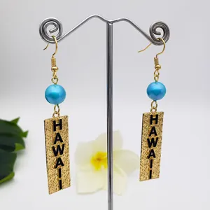 Detail of Hawaii themed gold drop earrings with blue bead and engraved metal plates