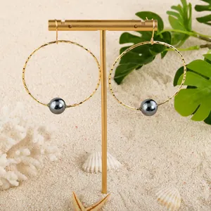 Golden hollow hoop earrings with large dark pearl and textured finish on beach background
