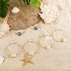 Three pairs of gold hoop earrings with pearl and shell charms on white beach sand