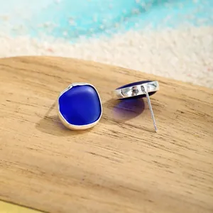 Detailansicht quadratischer, matter Meerglas-Ohrstecker in Blau auf Sandhintergrund