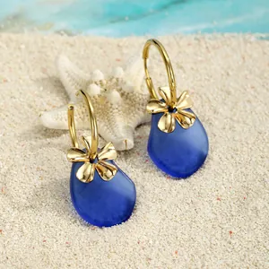 Detail von blauen Meeresglas-Ohrringen mit goldenen Blumen auf weißem Strandsand