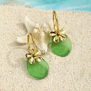 Schicke Seeglas-Ohrringe mit Goldblumen-Anhängern auf einem Sandstrand-Hintergrund