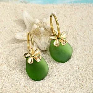 Nahaufnahme grüner Meeresglas-Ohrringe mit goldenen Blumen auf weißem Sandstrand
