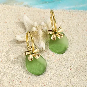 Detailansicht der Goldblumen-Ohrringe mit grünem Seeglas-Anhänger auf weißem Sand und Seestern