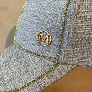 Detailed close-up of light blue tweed baseball cap with gold M logo and rhinestone chain trim