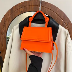 Minimalist bright orange square handbag with knotted handle and shoulder strap shown in hand