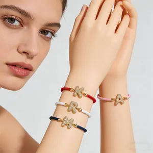 Detail von geflochtenen Armbändern mit vergoldeten Initialen-Anhängern aus Zirkonia-Mikropavé