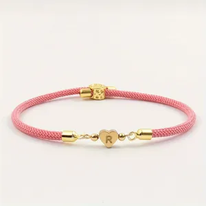 Detailansicht eines rosa geflochtenen Armbands mit goldenem Herzanhänger und Buchstaben R