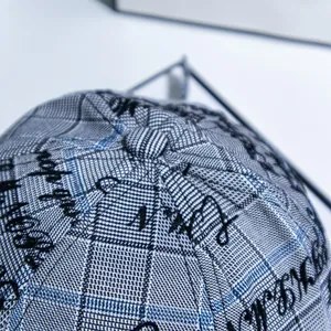 Detail shot of the baseball cap's top button and grey blue plaid fabric with black embroidery