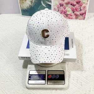 Dettaglio del cappello bianco con strass e logo C dorato su bilancia digitale che segna 91g