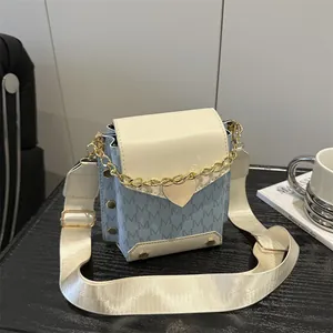 Bolso bandolera azul con monograma, solapa crema y cadena de corazón dorada sobre mesa