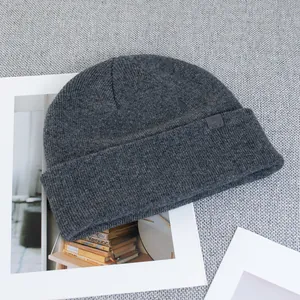 Detail shot of a dark grey wool knit beanie featuring a square logo patch on a textured surface