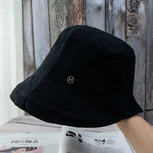 Detail of a black cotton bucket hat featuring a silver metal M logo and side pleats