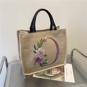 Beige Jute-Tragetasche mit Blumen-Buchstabe O und schwarzen Griffen auf Metallregal