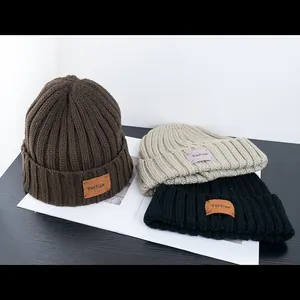 Three rib-knit beanie hats in dark brown, beige, and black with a leather logo patch