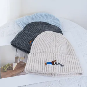 Détail de la broderie de teckel sur des bonnets en tricot bleu, gris et blanc crème