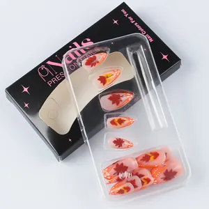Detail of nude press-on nails with maple leaf motifs and orange tips in premium packaging