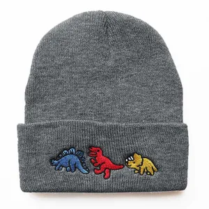Detailaufnahme einer grauen Strickmütze mit drei bunten Cartoon-Dinosaurier-Stickereien