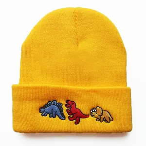 Nahaufnahme von drei Dinosaurier-Stickereien auf einer gelben Strickmütze für Damen