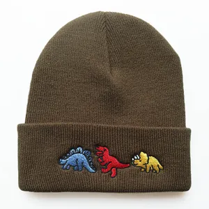Nahaufnahme der drei bunten Dino-Stickereien auf einer dicken braunen Strickmütze