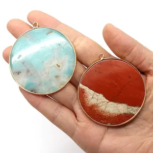 Hand holding round stone pendants with gold metal edges in blue and reddish-brown colors