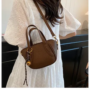 Details einer braunen Leder-Eimertasche mit Litschi-Textur und goldener Glocke am weißen Kleid