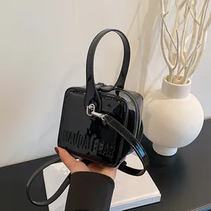 Detail of hand holding a black patent leather square box bag with gold-tone dual zippers