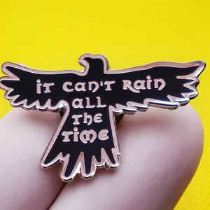 Black and gold raven enamel pin with 'It Can't Rain All The Time' text and spread wings