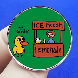 Round enamel pin featuring The Duck Song yellow duck and lemonade stand on green background