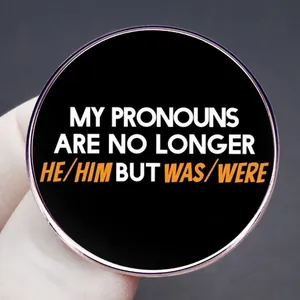 Round black enamel pin with My Pronouns Are No Longer He/Him But Was/Were text design