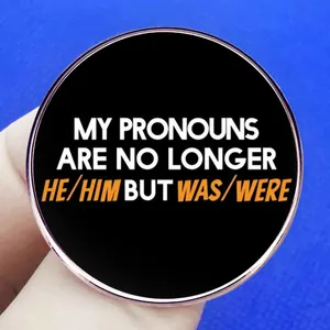Round Was/Were pronouns enamel pin badge held by hand against a blue background