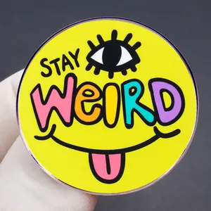 Gelber Stay Weird Emaille-Anstecker mit Auge und Smiley auf grauem Hintergrund