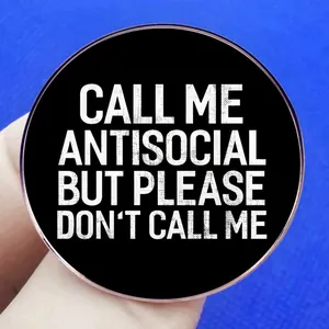 Round black enamel pin with white text 'Call me antisocial' held against blue fabric