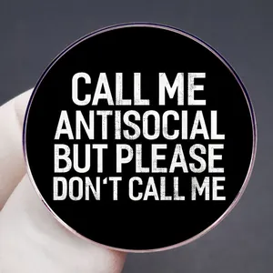 Round black enamel pin with white text Call Me Antisocial humor quote held by fingers