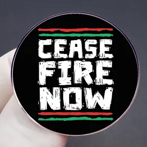 Cease Fire Now round metal badge pin with flag colors held by hand on grey background