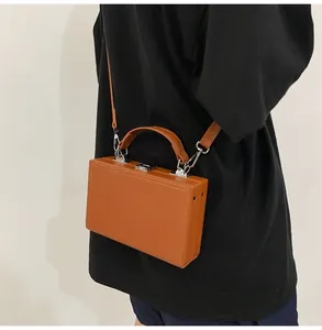Brown hard-shell box bag worn as a crossbody with top handle and silver buckle