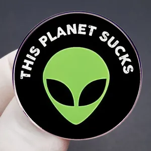 Round enamel pin featuring a green alien and This Planet Sucks quote on black metal