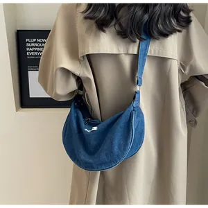 Detail shot of blue denim crescent bag with white woven label on a beige trench coat